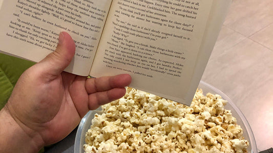 Hand holding a paperback book next to a tub of popcorn – perfect setup for a binge-worthy thriller.
